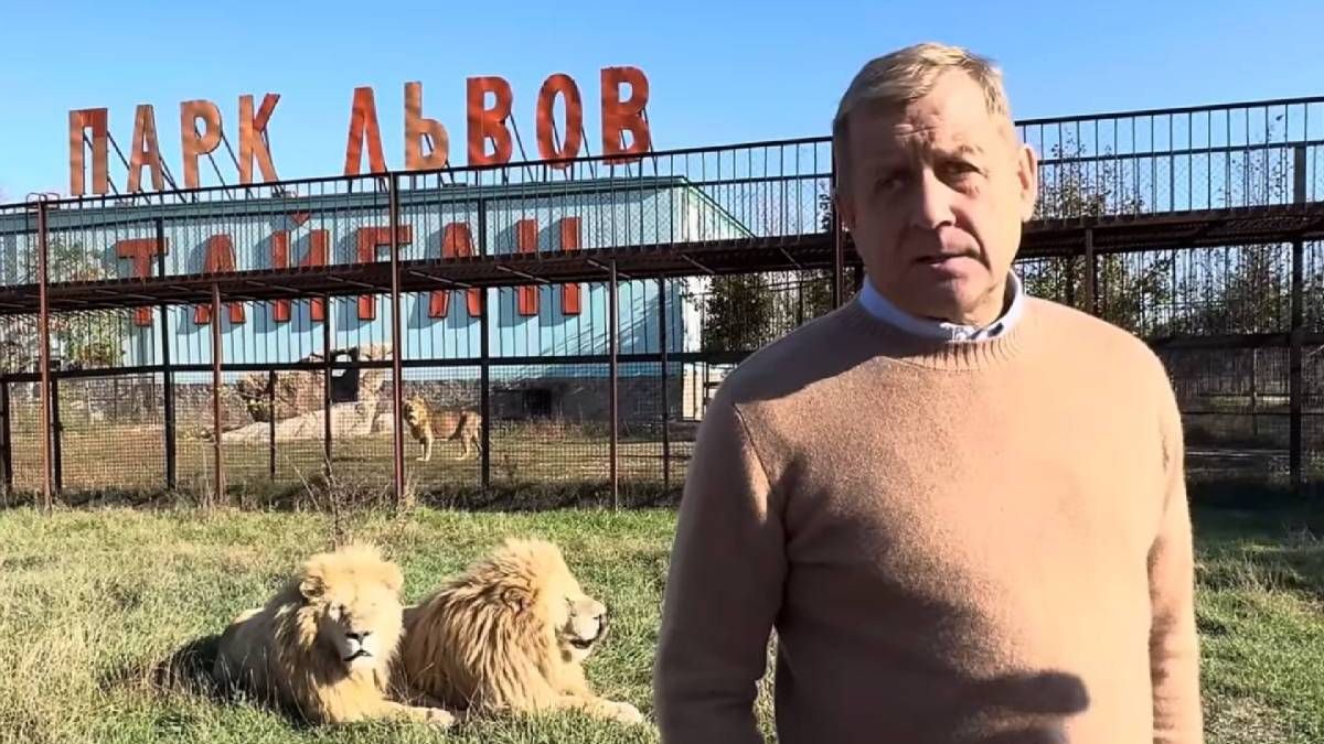 Чоловік у світлому светрі стоїть біля клітки з двома левами, ззаду напис українською \"ПАРК ЛЬВОВ ТАЙГАН\", сонячна погода.