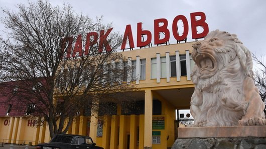 Жовта будівля з колонами, зверху великі червоні літери «ПАРК ЛЬВОВ». Праворуч велика світла кам’яна статуя лева на постаменті. Ліворуч дерево без листя і темний автомобіль. Небо сіре.