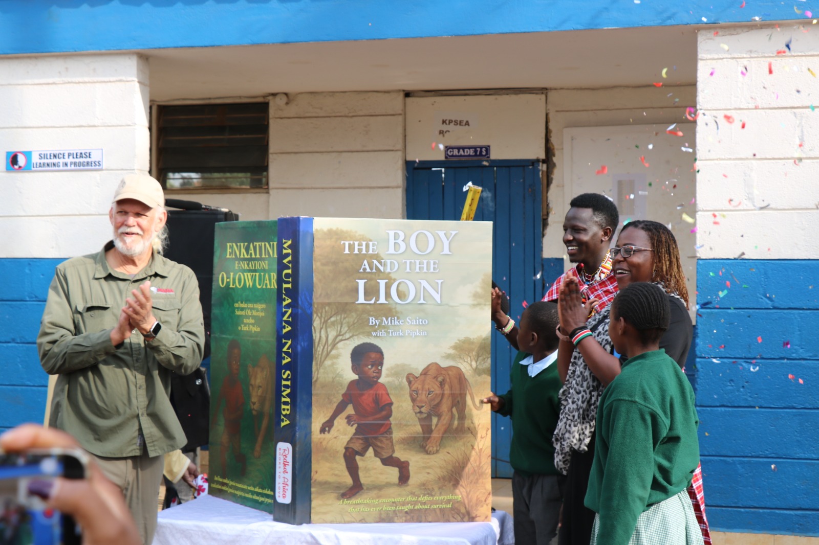 Люди радіють навколо великої книжки з написом «THE BOY AND THE LION». Стіни будівлі пофарбовані у синій та білий кольори. Помітні різнокольорові конфетті.